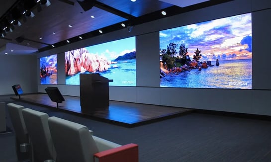 Auditorium with multiple large LED wall displays showing scenic ocean and mountain visuals behind a presentation stage.